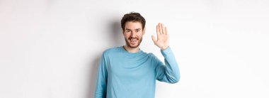 Sakallı neşeli genç adam merhaba diyor, dostça bakıyor ve el sallıyor, beyaz arka planda duruyor..