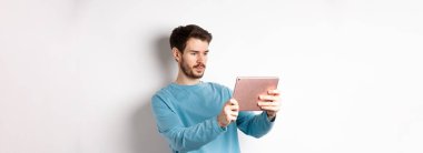 Genç adam dijital tablette fotoğraf çekiyor. Alet ekranına ciddi ciddi bakıyor. Beyaz arka planda duruyor..