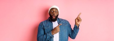 Harika bir tanıtım teklifi sunan siyahi adam, kameraya bakıyor, heyecanlı ve gülümsüyor, sol üst köşedeki afişi işaret ediyor, pembe arka plan..