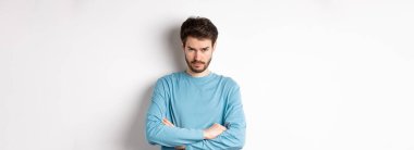 Kırgın ve üzgün sakallı genç bir adamın görüntüsü, alnının altından bak ve asık suratlı, kollarını göğsüne daya, kızgın, beyaz arka planda duran birine..