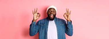 Bereli ve gömlekli etkileyici siyahi adam, tamam işaretleri gösteriyor ve kameraya şaşkın şaşkın bakıyor, gülümsüyor, harika bir teklif sunuyor, ürün öneriyor, pembe arka plan..