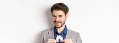 Gayrimenkul ve sigorta konsepti. Gülümseyen erkek emlakçı küçük kağıt ev resimleri gösteriyor ve kameraya bakıyor, mülk satıyor, beyaz arka plan.