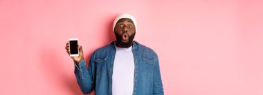 Sokak tarzı giyinmiş, cep telefonu ekranı gösteren, akıllı telefon görüntüsü sergileyen, pembe arka planda duran siyahi bir adam..