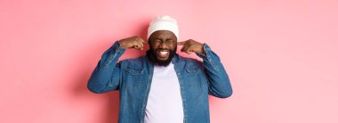 Memnun olmayan Afro-Amerikalı adam kulaklarını kapattı ve korkunç gürültüden yüzünü buruşturdu, rahatsız edici sesten rahatsız oldu, pembe arka planda durdu..