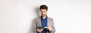 Çevrimiçi kredi kartı ve akıllı telefonla ödeme yapan yakışıklı bir işadamı, takım elbise içinde beyaz arka planda duruyor..