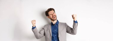 Takım elbiseli başarılı iş adamı, rahatlamış gülümseme ve yumruk pompalarıyla evet diye bağırıyor, başarıyı ve başarıyı kutluyor, beyaz arka planda duruyor..