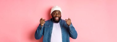 Mutlu Afro-Amerikalı hippi çocuk kamerayı işaret ediyor, sana ihtiyacı var, gülümsüyor ve şirkete davette bulunuyor, pembe arka planda duruyor..
