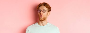 Kızıl saçlı, sakallı, kaşlarını kaldıran, şaşırmış ve pembe arka planda duran sağ üst logoya bakan meraklı genç adamın yüzü..