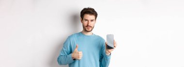 Boş akıllı telefon ekranı ve başparmağı gösteren genç adam, beyaz arka planda duran mobil uygulama veya çevrimiçi alışveriş önerisi.