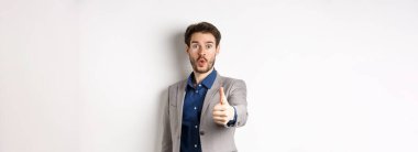 Takım elbiseli yakışıklı iş adamının görüntüsü. Heyecanlı yüzüyle baş parmağını gösteriyor. Beyaz arka planda duruyor..