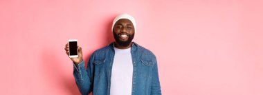 Bereli ve kot gömlekli yakışıklı hippi gülümsüyor, mutlu yüzüyle cep telefonu ekranını gösteriyor, uygulamayı tanıtıyor, pembe arka planda duruyor..