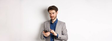 Yakışıklı sakallı erkek model cep telefonu kullanıyor, kameraya gülümsüyor, beyaz arkaplan.