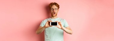 Şaşırmış kızıl saçlı adam kameraya hayranlıkla bakıyor, yatay olarak boş bir akıllı telefon ekranı gösteriyor, pembe arka planda duruyor..