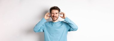 Neşeli adam kameraya göz kırpıyor ve gülümsüyor, kablosuz kulaklıkla müzik dinliyor, beyaz arka plan.