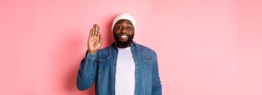 Dostça gülümseyen siyah adam merhaba diyor, el sallıyor, seni selamlıyor, pembe arka planda duruyor..