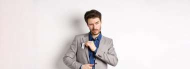 Kendine güvenen ve başarılı iş adamı bir kenara çekilirken kredi kartını takım elbisenin cebine koyup beyaz arka planda duruyor..