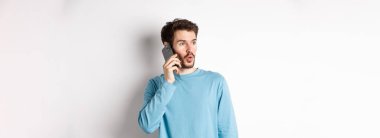 Teknoloji konsepti. İlgi çekici genç adam soldan etkilenmiş görünüyor, cep telefonuyla konuşuyor, birini arıyor, beyaz arka planda duruyor..