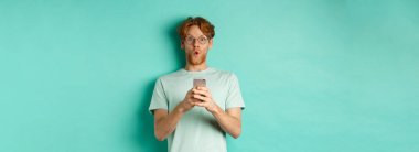 Kızıl saçlı, gözlüklü, elinde cep telefonu olan etkileyici bir genç adam kameraya hayret ediyor ve bakıyor..