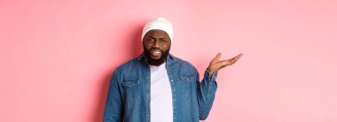 Sinirli ve kafası karışmış siyah adam tartışıyor, elini kaldırıyor ve kameraya şaşkın şaşkın bakıyor, ne olduğunu anlayamıyor, pembe arka planda duruyor..