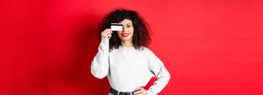 Kıvırcık saçlı, plastik kredi kartı gösteren, gülümseyen, kırmızı arka planda duran güzel bir manken. Makyaj ve alışveriş konsepti.