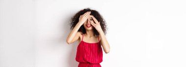 - Sürpriz. Heyecanlı, kıvırcık saçlı, kırmızı dudaklı, parmaklarının arasından bakan, şaşkın bir şekilde gülümseyen, tanıtıma bakan, beyaz arka planda duran güzel kız..