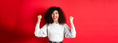 Kıvırcık saçlı neşeli şanslı kadın, ödül kazanıp sevinçle evet diye haykırıyor, ellerini kaldırıyor ve kutluyor, zafer ve tezahürat yapıyor, kırmızı arka planda duruyor..