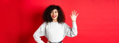 Heyecanlı kıvırcık saçlı kadın parmaklarıyla beş numarayı gösteriyor, düzen kuruyor, kırmızı arka planda duruyor..