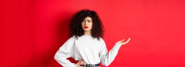 Kafası karışmış ve şüpheci bir kadın, kıvırcık saçlı, kaşlarını çatan ve el kaldıran, kırmızı arka planda duran garip bir şeyi anlayamıyor..