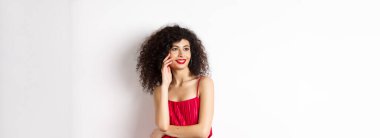 Kırmızı elbiseli ve makyajlı zarif ve romantik bir kadın. Gülümsüyor ve yüzüne dokunuyor. Mutlu bir ifadeyle, beyaz arka planda..