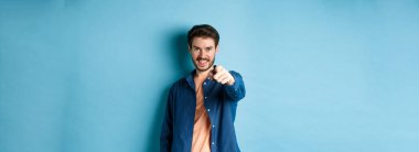 Kendinden emin gülümseyen adam seni seçiyor ya da davet ediyor, kamerayı işaret ediyor kararlı, mavi arka planda duruyor..