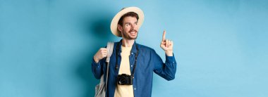 Tatilde yakışıklı, yaz şapkası takan, logoya bakıp işaret eden, gülümseyen, elinde kamera ve sırt çantasıyla mavi arka planda duran bir adam..