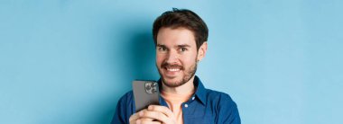 Sakallı, mutlu bir şekilde kameraya gülümseyen ve elinde cep telefonu olan mavi arka planda duran genç adama yakın durun..