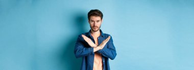 Ciddi beyaz adam hayır diyor, çapraz duruş hareketi yapıyor ve kameraya kendinden emin bakıyor, kötü bir şeyi yasaklıyor, mavi arka planda duruyor..