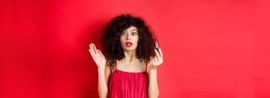 Güzellik ve makyaj konsepti. Kıvırcık saçlı heyecanlı bir kadın, kameraya bakarken nefesi kesiliyor, kırmızı ruj tutuyor, beyaz arka planda elbise giyiyor..