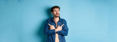 Kararsız sakallı adam çapraz kollar ve iki farklı türü gösteren, seçim yapan, mavi arka planda duran.