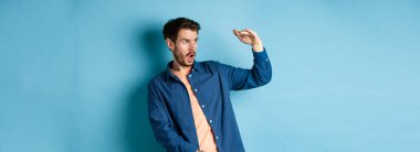 Büyük bir nesneyle boş alana bakan, büyük boyutlarda, mavi arka planda duran, etkileyici bir beyaz adam. Boşluğu kopyala