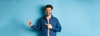 Gülümseyen, kendine güvenen, logo gösteren, boş alanı gösteren ve mavi arka planda duran, kameraya bakan bir adam..