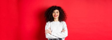Makyajlı, kıvırcık saçlı, göğsünde çapraz kolları olan ve profesyonel görünümlü, kırmızı arka planı olan kendine güvenen bir kadın..