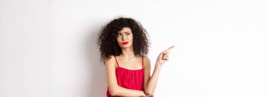 Kıvırcık saçlı, kırmızı elbiseli, solda logoyu gösteren, beyaz arka planda duran, hayal kırıklığına uğramış ve şüpheci genç bir kadın..