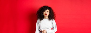 Kadın hasta, eğiliyor ve karnına dokunuyor, karın ağrısı ya da regl sancısı çekiyor, kırmızı arka planda duruyor..