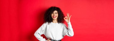 Kendine güvenen güler yüzlü kadın, onaylamak ya da ürünleri sevmek için onay işareti gösteriyor, iyi kalite garanti ediyor, iltifat ediyor, kırmızı arka plan.