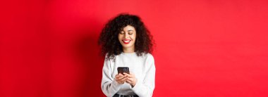 Mutlu pozitif kız telefonda konuşuyor, mesaj okuyor ve gülümsüyor, sosyal medya uygulamasını kullanıyor, kırmızı arka planda duruyor..