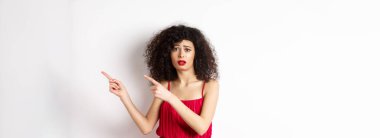 Şaşkın ve üzgün, kıvırcık saçlı, gece elbisesi ve makyajı olan, parmaklarını sağa doğru gösteren, üzgün, beyaz arka planda duran bir kadın..