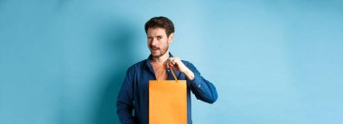 Turuncu alışveriş torbası gösteren, göz kırpan, mağaza indirimi öneren, mavi arka planda duran mutlu genç adam..
