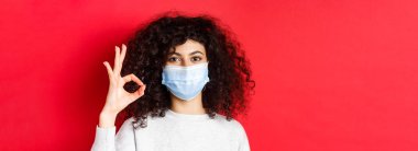 Covid ve sağlık konsepti. Tıbbi maskeli kendine güvenen genç bir kadın onay veren, kalite garantili, kırmızı arka planda duran bir jest gösteriyor..