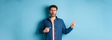 Şok olmuş ve gücenmiş genç adam kaşlarını çatmış, yaralı görünüyor, boş alanı işaret ediyor, bir şeyden şikayet ediyor, mavi arka planda duruyor..