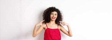 Kıvırcık saçlı, makyajlı, kırmızı elbiseli, kendini gösteren, beyaz dişli, beyaz arka planda duran güzel bir kadın..