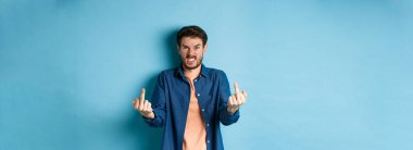 Angry rude guy showing middle finger and saying fuck you, swearing and staring furious at camera, standing on blue background.