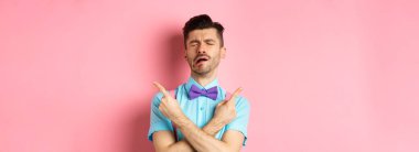 Papyonlu, üzgün ve hayal kırıklığına uğramış genç bir adam, ağlayan ve parmaklarını yana doğru gösteren, iki şey gösteren, karşılıksız aşktan ağlayan, pembe arka planda duran.