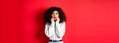 Şaşırmış beyaz kadın kıvırcık saçlı, etkileyici bir şeye bakıyor, hayrete düşmüş, reklam teklifini kontrol ediyor, kırmızı arka plan.